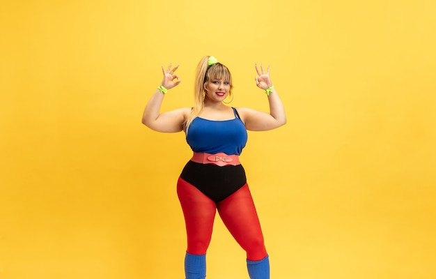 Jonge Kaukasische plus size vrouwelijke model opleiding op gele muur. Stijlvolle vrouw in lichte kleding. Kopieerruimte. Concept van sport, gezonde levensstijl, lichaamspositief, mode. Glimlachen, aardig tonen.