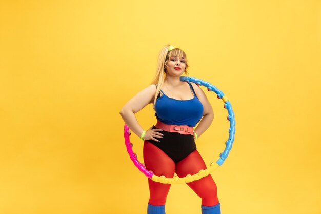 Jonge Kaukasische plus size vrouwelijke model opleiding op gele muur. Stijlvolle vrouw in lichte kleding. Kopieerruimte. Concept van sport, gezonde levensstijl, lichaamspositief, mode. Poseren met de hoepel.