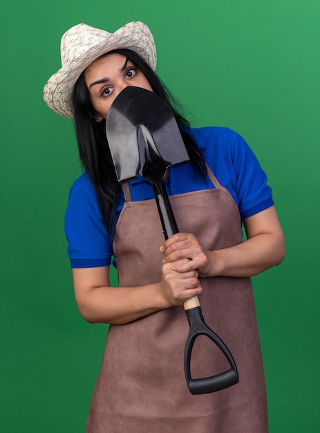 Jonge kaukasische tuinmanvrouw die uniform en hoed draagt die schop van erachter houden