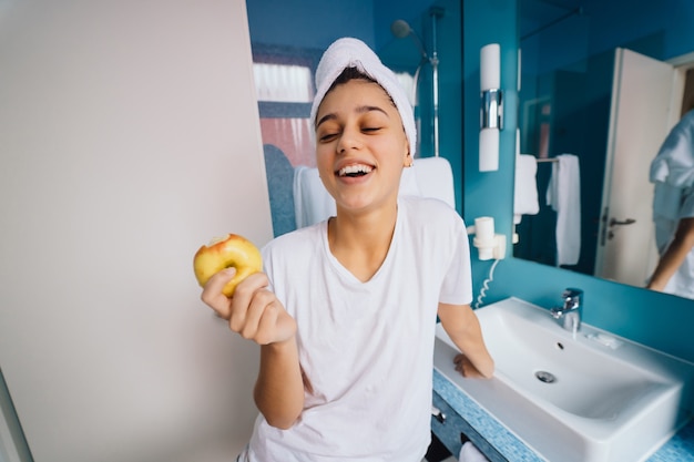 Jonge kaukasische vrouw die handdoek op hoofd en t-shirt in de appel van de badkamersholding draagt.