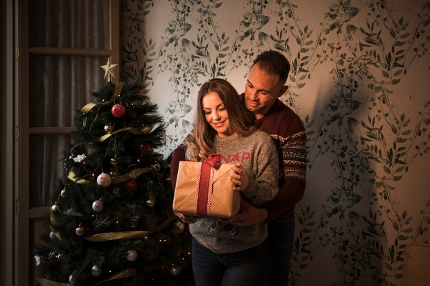 Jonge kerel die van achter vrolijke dame koestert met giftdoos dichtbij Kerstboom