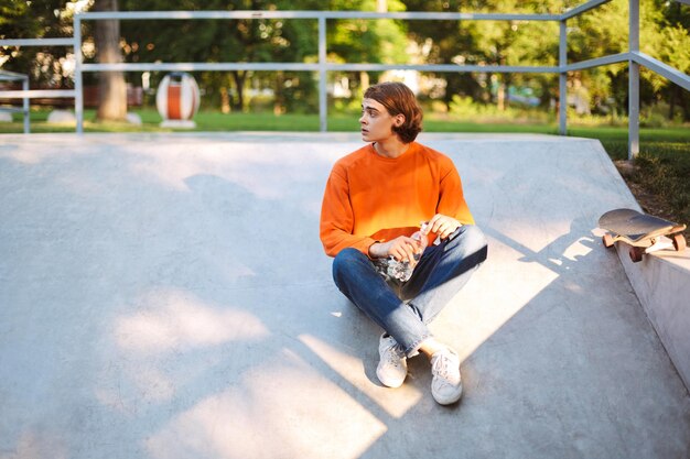 Jonge kerel in oranje trui en spijkerbroek met fles water in de hand terwijl hij bedachtzaam opzij kijkt naar skatepark