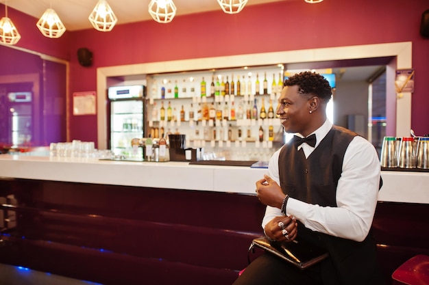 Jonge knappe afrikaanse man met een wit overhemd, een zwart vest en een vlinderdas met een portemonnee die tegen de bar in de nachtclub staat?