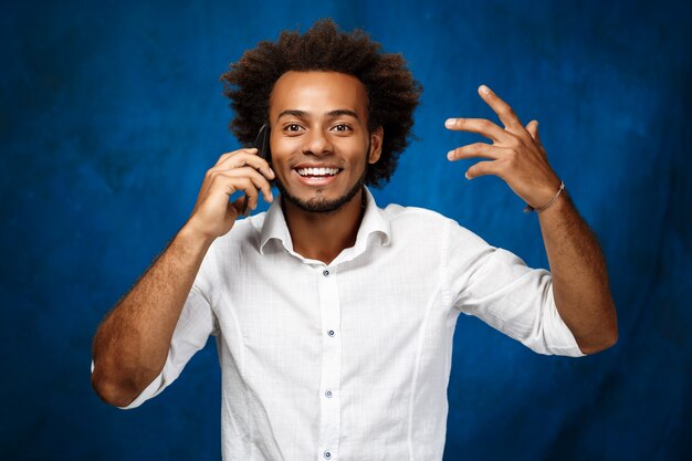 Jonge knappe Afrikaanse mens die op telefoon over blauwe muur spreekt.
