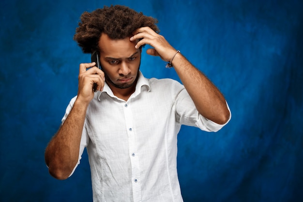 Jonge knappe Afrikaanse mens die op telefoon over blauwe muur spreekt.