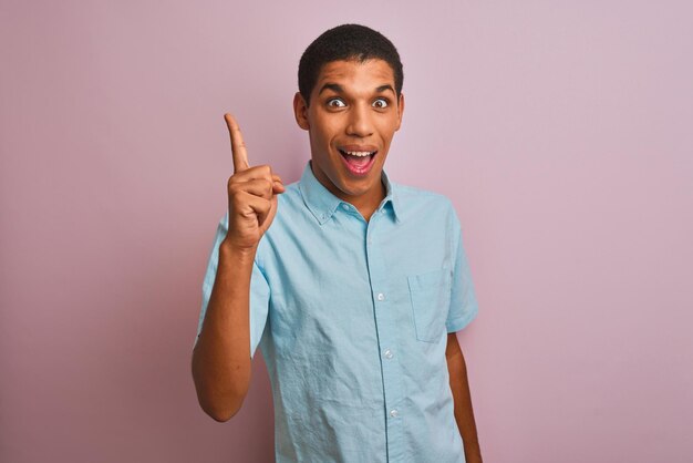 Jonge knappe arabische man met blauw shirt die over een geïsoleerde roze achtergrond staat en met de vinger omhoog wijst met een succesvol idee Verlaten en gelukkige nummer één