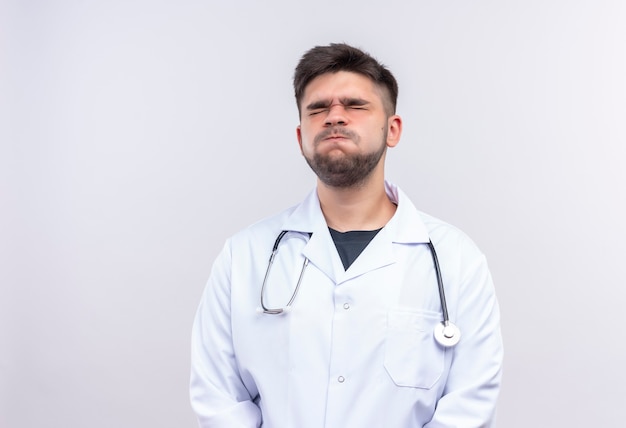 Jonge knappe arts die witte medische toga draagt, witte medische handschoenen en een stethoscoop die de pijn verdragen sluitende ogen die zich over witte muur bevinden