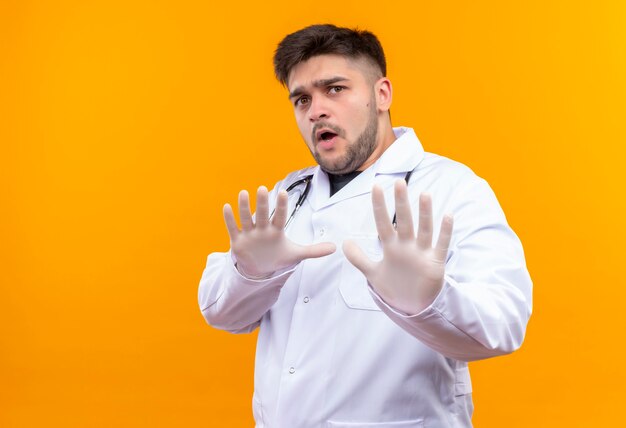 Jonge knappe arts die witte medische toga draagt, witte medische handschoenen en stethoscoop bang ophouden met handen die zich over oranje muur bevinden