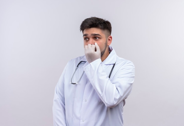 Jonge knappe arts die witte medische toga draagt, witte medische handschoenen en stethoscoop kijkt bang naast het houden van vuist op muis die zich over witte muur bevindt