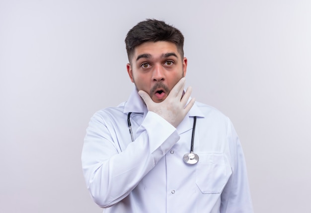 Jonge knappe arts die witte medische toga draagt, witte medische handschoenen en stethoscoop kijkt verbaasd terwijl hij zijn kaak met hand houdt die zich over witte muur bevindt