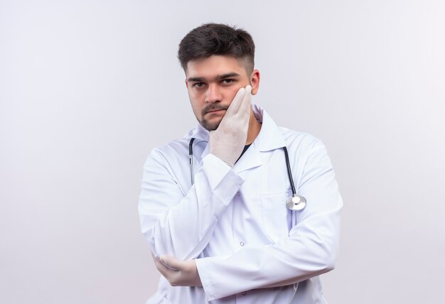 Jonge knappe arts die witte medische toga, witte medische handschoenen en een stethoscoop draagt die bedachtzaam zich over witte muur bevindt