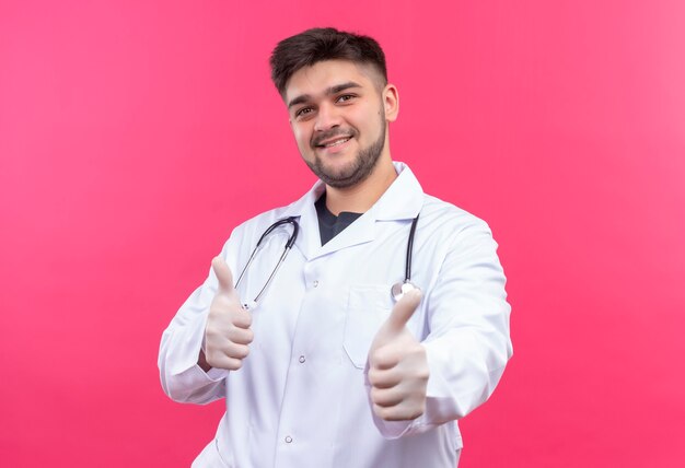Jonge knappe arts die witte medische toga witte medische handschoenen en stethoscoop draagt die gelukkige duimen omhoog doen die zich over roze muur bevinden