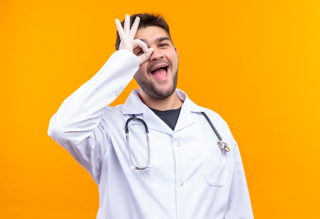 Jonge knappe arts die witte medische toga witte medische handschoenen en stethoscoop draagt die ok teken op zijn oog met hand doen die zich over oranje muur bevindt