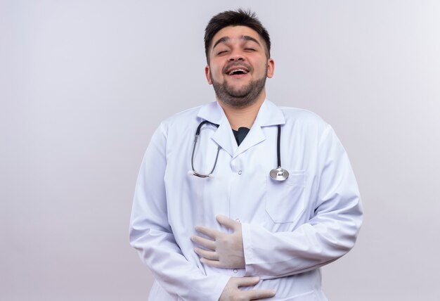 Jonge knappe arts die witte medische toga, witte medische handschoenen en stethoscoop draagt die zich over witte muur bevinden lachen