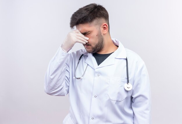 Jonge knappe arts die witte medische toga witte medische handschoenen en stethoscoop draagt die zijn lopende neus sluit die zich over witte muur bevindt