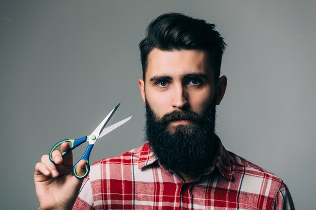 Jonge knappe bebaarde man met lange baard, snor en donkerbruin haar met kapper of kapper schaar met emotioneel gezicht op grijze muur