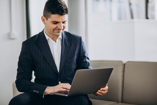 Jonge knappe bedrijfsmens die aan computer op een bank in bureau werkt