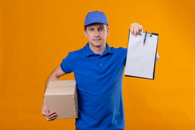 Jonge knappe bezorger in blauw uniform en pet met doos pakket en klembord met zelfverzekerde serieuze uitputting wachtend op handtekening staande over oranje muur