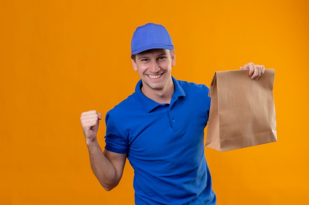 Jonge knappe bezorger in blauw uniform en pet met pizzadozen vuist heffen blij en tevreden verheugend zijn succes winnaar concept staande over oranje muur