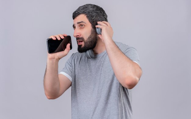 Jonge knappe blanke man met koptelefoon doen alsof zingen en zijn mobiele telefoon gebruiken als microfoon en kijken naar kant met hand op hoofdtelefoon geïsoleerd op een witte achtergrond met kopie ruimte