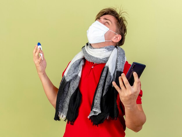 Jonge knappe blonde zieke man met masker en sjaal met thermometer en mobiele telefoon opzoeken geïsoleerd op olijfgroene achtergrond