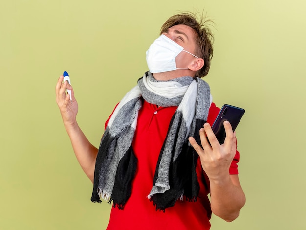 Jonge knappe blonde zieke man met masker en sjaal met thermometer en mobiele telefoon opzoeken geïsoleerd op olijfgroene achtergrond