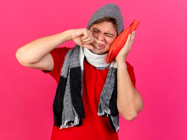 Jonge knappe blonde zieke man met winter hoed en sjaal aanraken van gezicht met warmwaterkruik neus afvegen met vinger met gesloten ogen geïsoleerd op crimson achtergrond met kopie ruimte