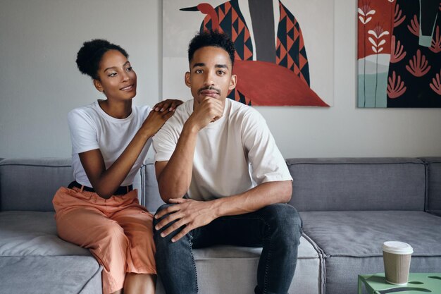 Jonge knappe, casual Afro-Amerikaanse man die bedachtzaam in de camera kijkt terwijl zijn vriendin hem dromerig aankijkt op de bank in een modern huis