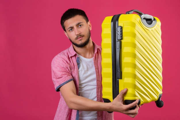 Jonge knappe de holdingskoffer die van de reizigerskerel camera met zekere ernstige uitdrukking op gezicht bekijken die zich over roze achtergrond bevinden