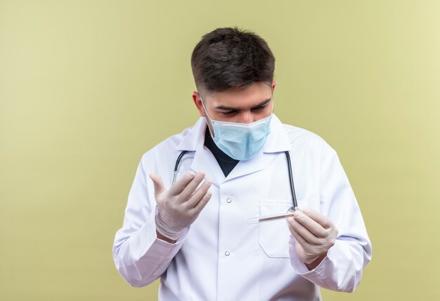 Jonge knappe dokter die een blauw medisch masker draagt, een witte medische jurk, een witte medische handschoenen en een stethoscoop die een thermometer houdt die geschokt is vanwege de temperatuur die over de kaki muur staat