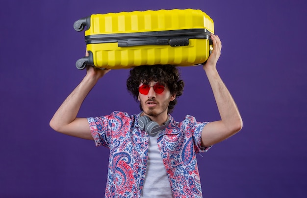 Jonge knappe gekrulde reiziger man met zonnebril en koptelefoon op de nek met koffer op het hoofd op geïsoleerde paarse muur