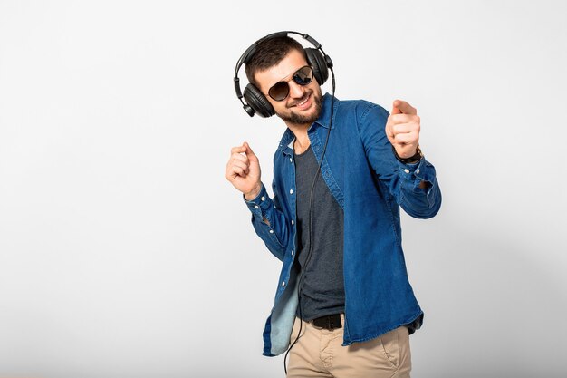 Jonge knappe gelukkig lachende man dansen en luisteren naar muziek in koptelefoon geïsoleerd op witte studio muur