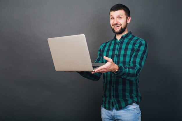 Jonge knappe hipster bebaarde man met laptop in handen, groen geruit overhemd, positieve emotie, gelukkig, glimlachen, verrassing, grijze achtergrond