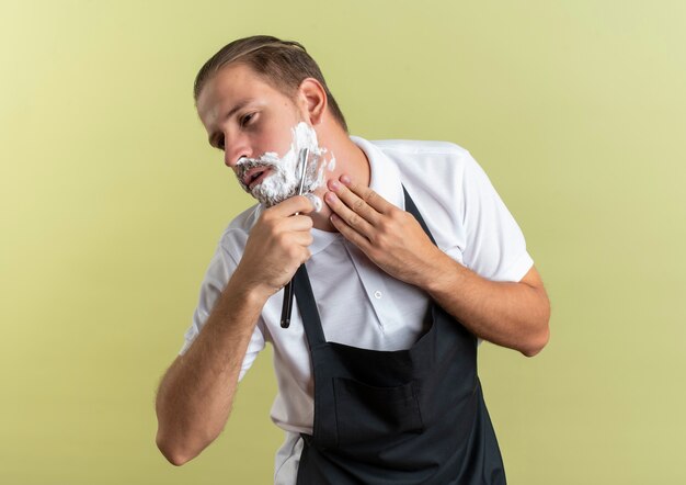 Jonge knappe kapper dragen uniform zijn baard scheren met een scheermes en raken zijn nek kijken kant geïsoleerd op olijfgroene achtergrond