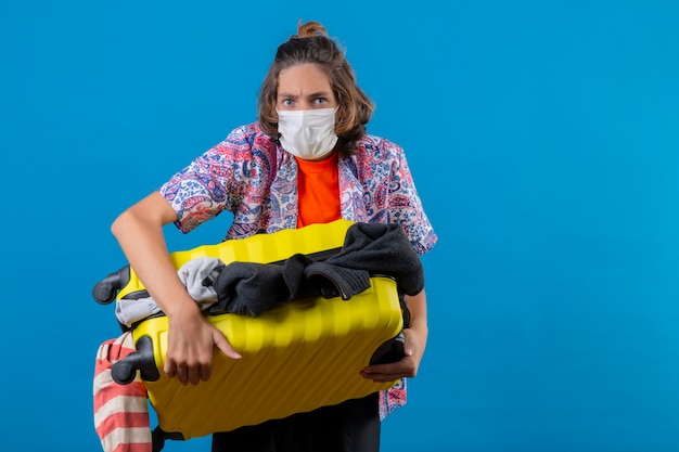 Jonge knappe kerel die een beschermend gezichtsmasker draagt en een reiskoffer vol kleren houdt die verward kijkt en geen antwoord heeft wat te doen staande over de blauwe achtergrond