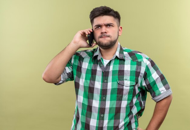 Jonge knappe kerel die een geruit overhemd draagt dat boos op telefoon spreekt die zich over kaki muur bevindt