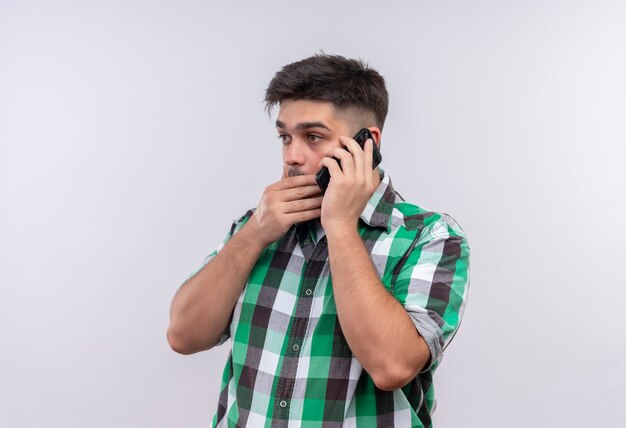 Jonge knappe kerel die een geruit overhemd draagt dat over telefoon spreekt geschokt kijkt naast status over witte muur