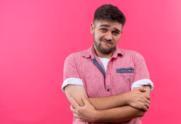 Jonge knappe kerel die een roze poloshirt draagt en onbeslist kijkt die verlegen over roze muur staat