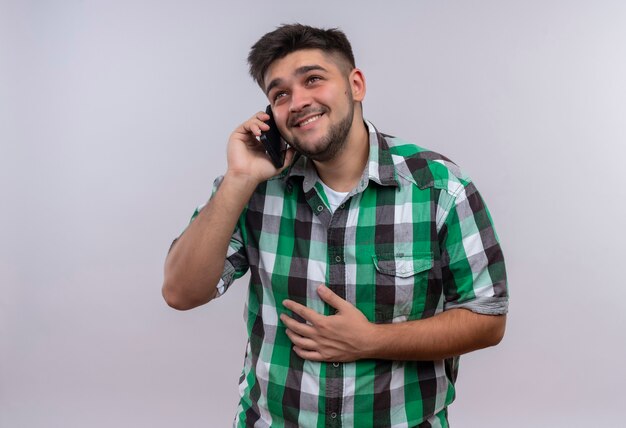 Jonge knappe kerel die geruit overhemd draagt dat en op telefoon lacht die zich over witte muur bevindt