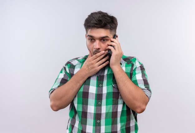 Jonge knappe kerel die geruit overhemd draagt geschokt sprekend over telefoon die zich over witte muur bevindt