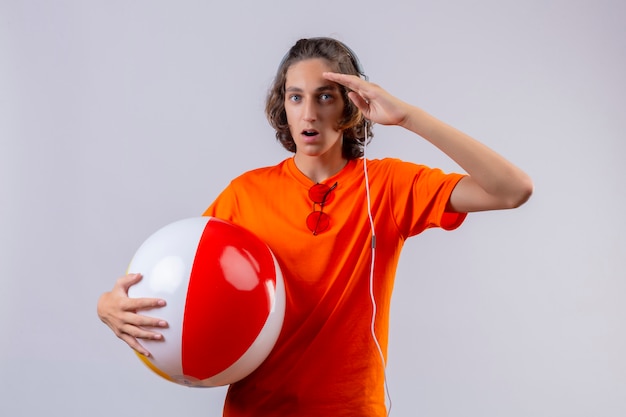 Jonge knappe kerel die in oranje t-shirt opblaasbare bal met hoofdtelefoons houden die verrast status met hand dichtbij hoofd kijken
