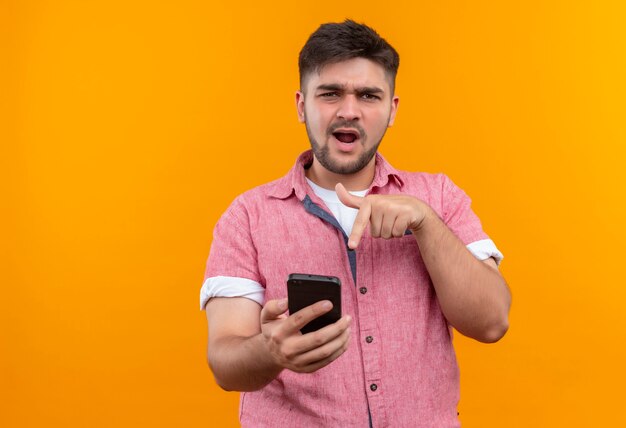 Jonge knappe kerel die roze polo draagt die naar telefoonoverhemd richt dat zich over oranje muur bevindt