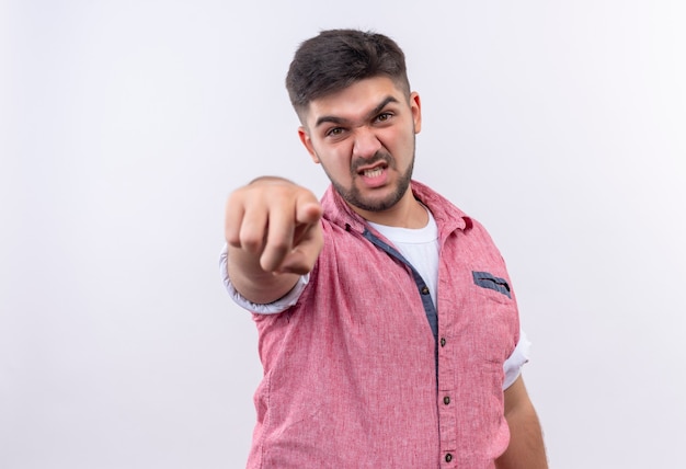 Jonge knappe kerel die roze poloshirt draagt dat boos naar camera richt die zich over witte muur bevindt