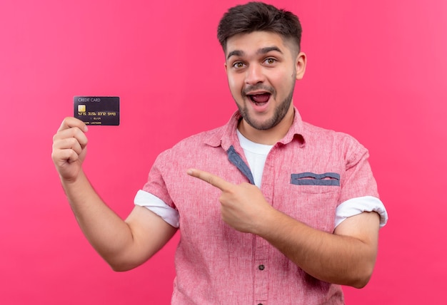 Jonge knappe kerel die roze poloshirt draagt dat gelukkig naar creditcard wijst die zich over roze muur bevindt