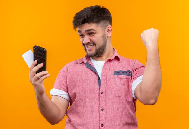 Jonge knappe kerel die roze poloshirt draagt dat telefoon en kaart bekijkt die zich in overwinning verheugt die zich over oranje muur bevindt