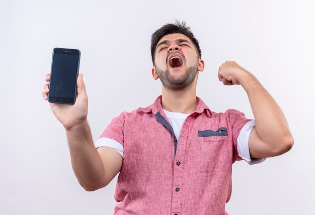 Jonge knappe kerel die roze poloshirt draagt dat zijn vuist schudt die telefoon sluitende ogen houdt die zich over witte muur bevinden