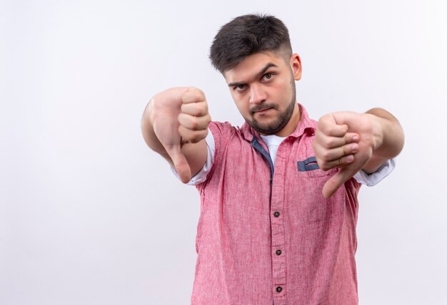 Jonge knappe kerel die roze poloshirt draagt die boos ongelukkig duimen doet die zich over witte muur bevinden