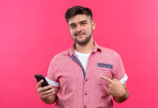 Jonge knappe kerel die roze poloshirt draagt die gelukkig naar de telefoon kijkt die zich over roze muur bevindt