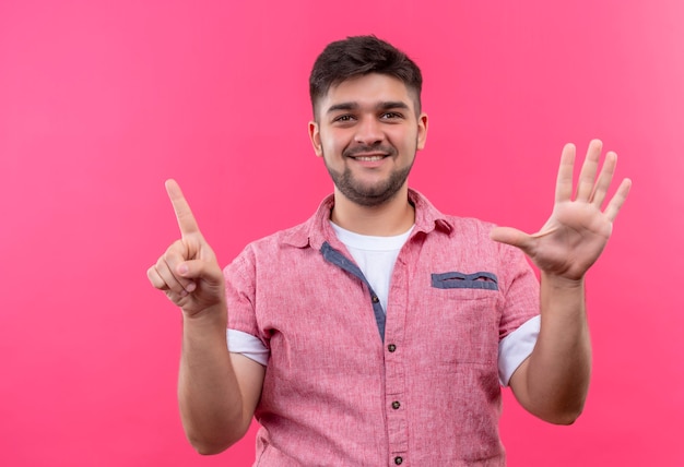 Jonge knappe kerel die roze poloshirt draagt die gelukkig zes met vingers toont die zich over roze muur bevinden