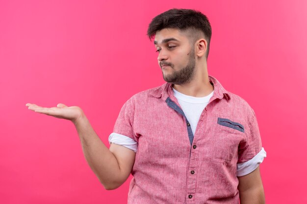 Jonge knappe kerel die roze poloshirt draagt die iets in zijn linkerhand houdt die zich over roze muur bevindt